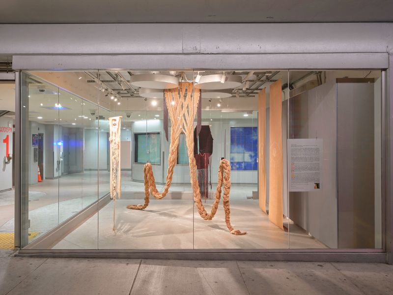 Fiber art installation with large braided ropes and textiles in a glass-fronted gallery.