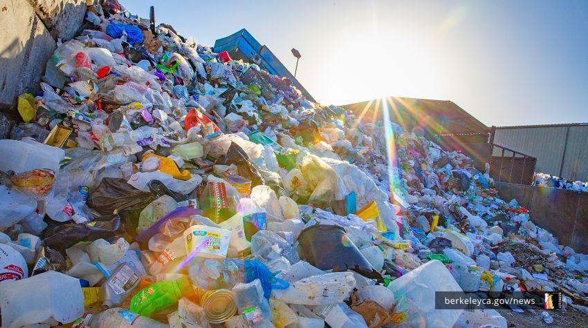Large pile of plastic and other waste materials at a recycling facility, with sunlight shining over the top of the heap.