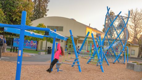 Child playing on blue climbing equipment at Grove Park playground near the recreation center, with the tall rope tower behind them.