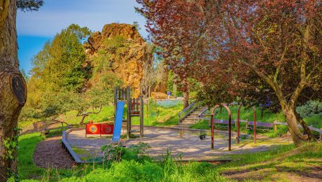 Playground area shaded by trees with climbing structures and a view of the large rock at Remillard Park in Berkeley.