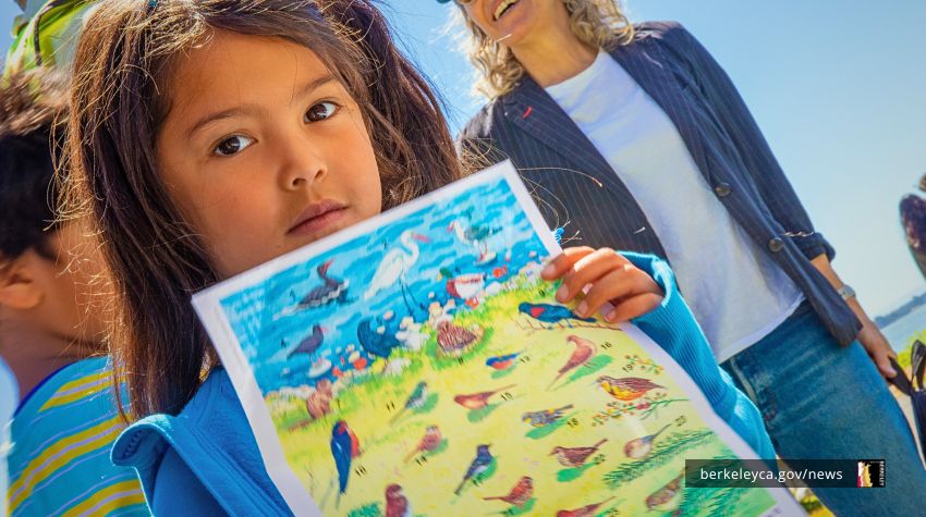 A young child looks at camera and holds up a colorful drawing of birds and ocean life while standing outside on a sunny day. An adult smiles in the background, and the Bay is visible in the distance.