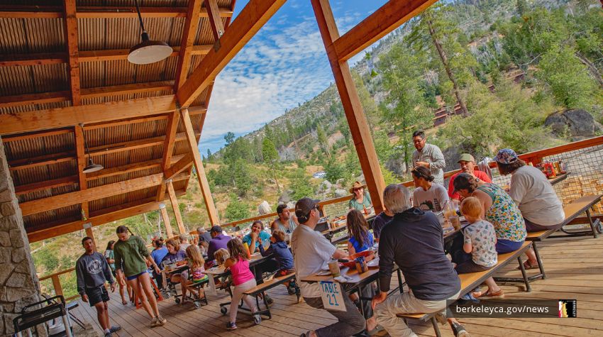 A group of families and children sit at wooden picnic tables under a large open-air pavilion in the mountains. People are eating, talking, and enjoying the forested outdoor setting. The tables overlook a scenic landscape of trees and rocky hills.
