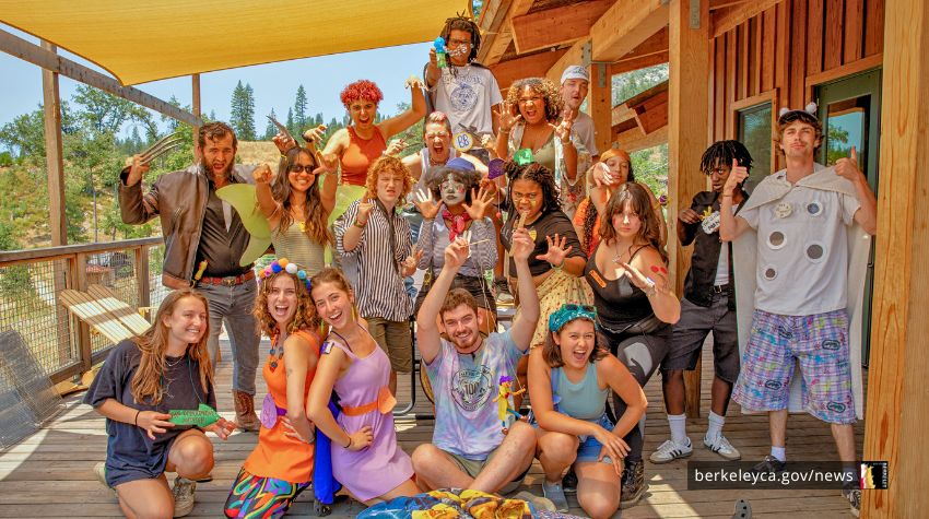 A large group of camp staff members pose together on a wooden deck outside a cabin, wearing colorful costumes and raising their hands while smiling at the camera.