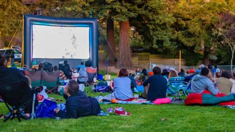 crowd sits on blankets and chairs in front of inflatable screen in a park